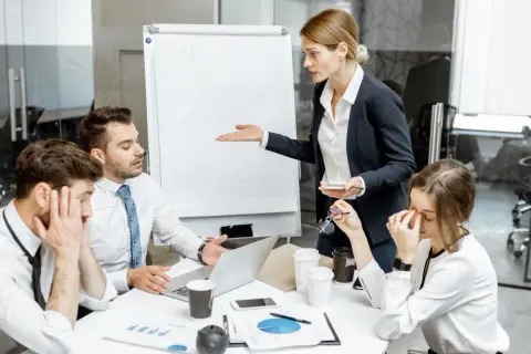 En kvinna gestikulerar mot en whiteboard medan kollegor ser utmattade ut. Känslan av manipulativt beteende hänger i luften under mötet.