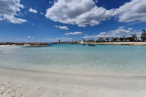 Kristallklart vatten och vit sand vid Nissi Beach, Cypern. Små båtar guppar i det turkosa vattnet under en blå himmel med vita moln.