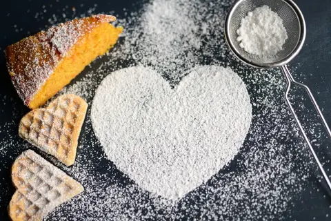 A slice of cake, heart-shaped cookies, and a heart shape made of powdered sugar on a dark surface.
