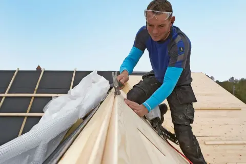 A roofer wearing safety glasses hammers a wooden plank onto a roof under construction.