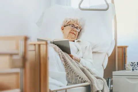 Ältere Dame liest im Bett, eine Auszeit in der Kurzzeitpflege ohne Pflegegrad. Gemütlich eingekuschelt mit einem Buch.