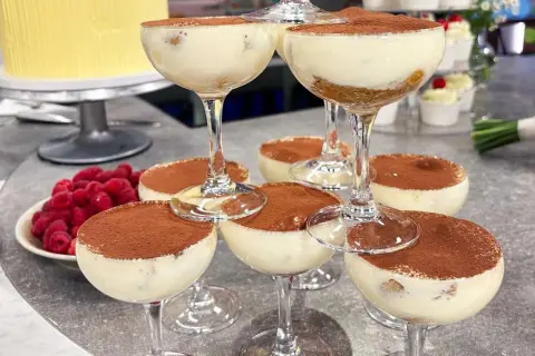 Flera glas med krämig tiramisu, pudrad med kakao. Hallon och en gul tårta i bakgrunden.