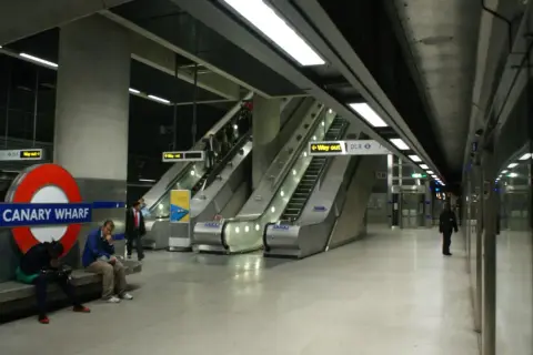 Stacja Canary Wharf w **metro londyn** z charakterystycznym znakiem, ruchomymi schodami i ludźmi czekającymi na pociąg.