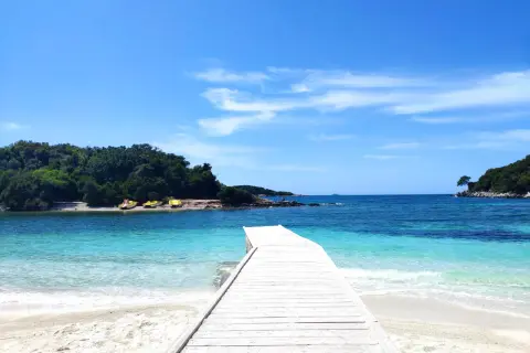 Białe molo prowadzi do krystalicznie czystej wody na Ksamil Beach. W oddali zielone wyspy i błękitne niebo.