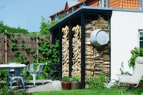 Vedbod med snyggt staplad ved. En utomhusplats med bord, stolar och en grill.