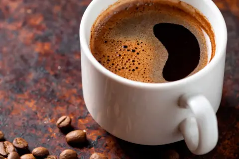 En vit kopp med mörkt kaffe och skum. Kaffebönor ligger utspridda. Undrar du hur mycket koffein i en kopp kaffe?