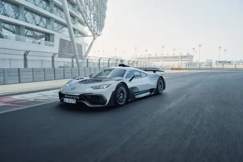 Srebrny Mercedes-AMG Project One pędzi po torze wyścigowym, obok nowoczesnej architektury.