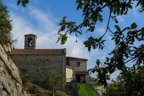 L'eremo di Cerbaiolo, con la sua torre campanaria in pietra, si staglia contro un cielo azzurro tra i rami di una quercia.