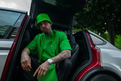 Malik Montana w zielonym kapeluszu i koszulce, siedzi w samochodzie z otwartymi drzwiami. Wygląda jak adwokat diabła, gotowy na nowe wyzwania.