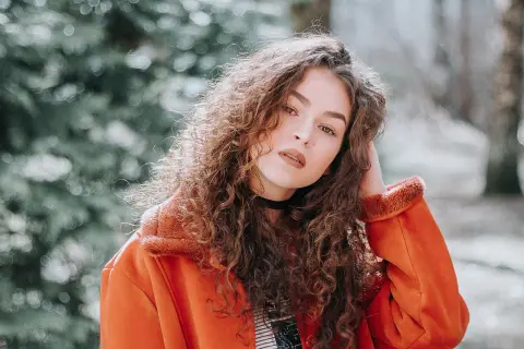 Joven con melenas rizadas y un abrigo naranja, posando con confianza.