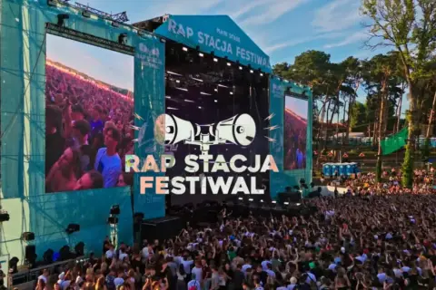 Tłum ludzi na koncercie rap festiwal. Na scenie wielkie ekrany z napisem "RAP STACJA FESTIWAL" i grafiką megafonów.