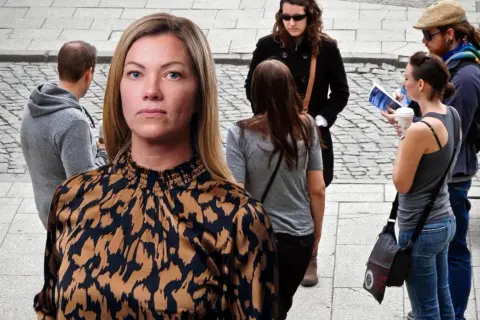 En kvinna med en mönstrad blus tittar framåt. I bakgrunden syns flera personer, en del verkar diskutera, vilket kan symbolisera ansvarstagande på jobbet.