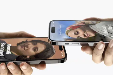 Dwie dłonie trzymają iPhone'y z funkcją udostępniania kontaktów w iOS 17. Na ekranach widnieją zdjęcia i dane Sonali Sanghvi oraz Athirah Abdullah.