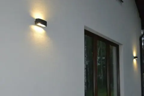 Dwa kinkietowe oświetlenie zewnętrzne rzuca ciepłe światło na białą, teksturowaną elewację, uwydatniając nierówności na elewacji pod światło.