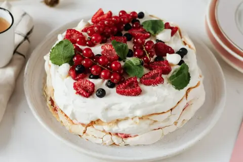 Pavlova z kremem do bezy, ozdobiona świeżymi malinami, borówkami i porzeczkami.