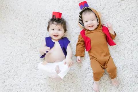 Dwa uśmiechnięte niemowlaki w strojach inspirowanych Alladynem leżą na białym dywanie. Jeden w fioletowej kamizelce, drugi w brązowym kombinezonie.