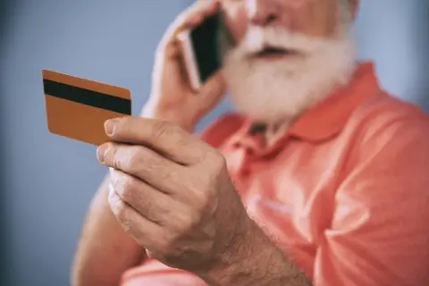 Starszy mężczyzna z brodą trzyma kartę i rozmawia przez telefon, zastanawiając się, dlaczego bank zablokował kartę.