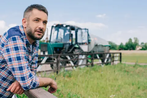 Rolnik w kratę oparty o płot, w tle traktor. Wsparcie dla małych gospodarstw to klucz do rozwoju wsi.