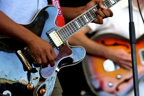 Hände spielen auf einer schwarzen E-Gitarre, während im Hintergrund eine weitere Gitarre zu sehen ist. Die Band probt für ihren nächsten Auftritt.
