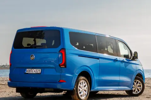 Błękitny VW Transporter T7 na piaszczystej plaży, z widokiem na morze.