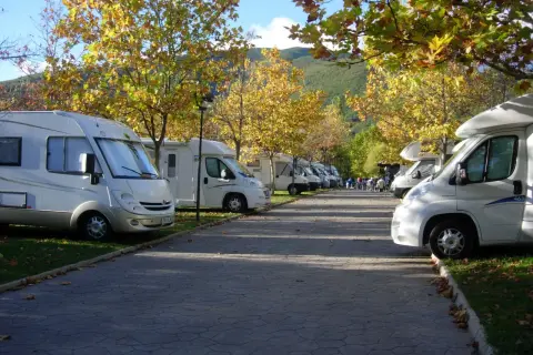 Un tranquilo caravan parking bajo árboles otoñales. Varias autocaravanas blancas aparcadas en fila, con hojas doradas cayendo.