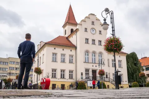 Mężczyzna patrzy na ratusz w Trzebnicy, jedno z wielu miejsc wartych zobaczenia. W tle zabytkowa architektura i kwitnące kwiaty.