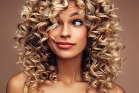 Kobieta z burzą blond loków, loki krótkie kręcone włosy tworzą ramę dla jej twarzy.