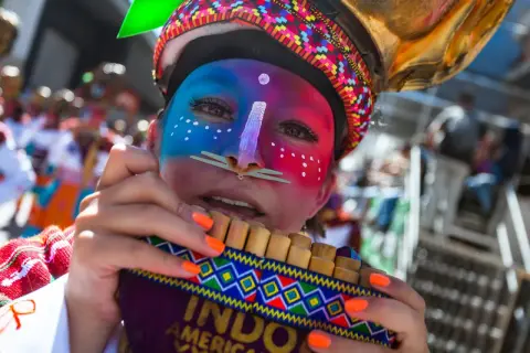 Młoda kobieta w barwnym makijażu i tradycyjnym nakryciu głowy gra na fletni Pana podczas parady latino.