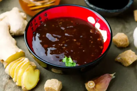 Sos teriyaki przepis: miseczka z ciemnym sosem, obok imbir, czosnek, brązowy cukier i miód.