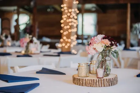 Un bouquet de fleurs roses et crème sur une tranche de bois, entouré de bougies dorées. Le **protocole fiançailles** est respecté avec des serviettes bleues sur des tables blanches, sous des guirlandes lumineuses.
