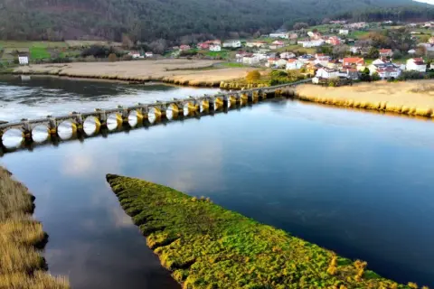 Un puente de piedra con arcos cruza un río azul, conectando un pueblo pintoresco con campos verdes. El puente, conocido como Ponte nafonso, se alza majestuoso sobre el agua.