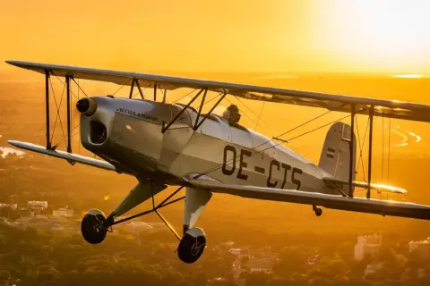 Ein silberner Doppeldecker fliegt im goldenen Abendlicht über einer Stadt. Ein Traum für alle, die gerne flugzeuge fotografieren.