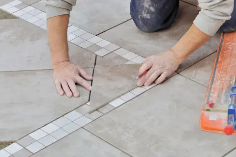 Pose en quinconce de carreaux de sol, avec une bordure décorative. Un carreleur marque une pièce avec un crayon.