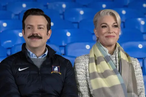 Die Besetzung von Ted Lasso, Jason Sudeikis und Hannah Waddingham, sitzt lächelnd auf blauen Tribünen.
