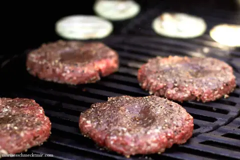 Vier rohe Burger-Patties, die darauf warten, dass man sie selber macht, brutzeln auf einem Grill. Zwiebelringe liegen im Hintergrund.
