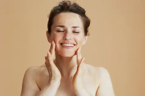 Kobieta z zamkniętymi oczami nakłada peeling na twarz, ciesząc się zabiegiem. Zastanawiasz się, jak często robić peeling twarzy?