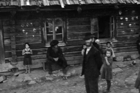 Kinder und Erwachsene stehen vor einem Holzhaus. Die Szene erinnert an die **Judenverfolgung** und die damit verbundene Not.