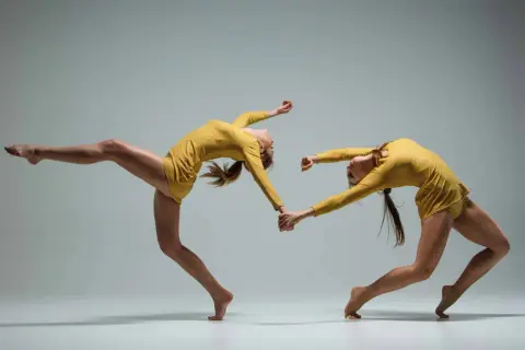 Deux danseuses en tenue jaune, mains jointes, en mouvement synchronisé sur fond gris.