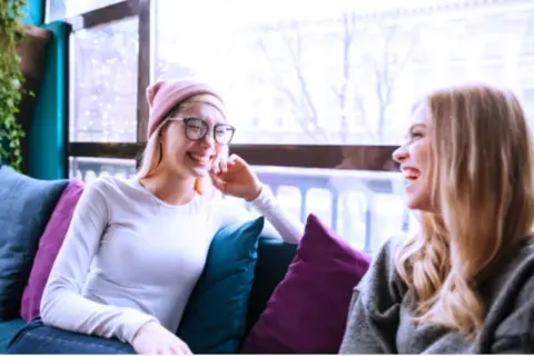 Dos amigas ríen sentadas en un sofá, como si compartieran anécdotas de couchsurfing que es una forma de viajar.