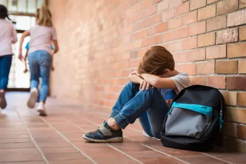 Chłopiec siedzi przy ceglanej ścianie, przytulając kolana. Inne dzieci biegną obok, nie zwracając na niego uwagi. To obraz samotności i fobia społeczna u nastolatków.