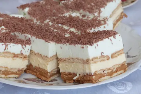 Szybkie ciasto bez pieczenia z budyniem i herbatnikami, przełożone kremem i posypane wiórkami czekoladowymi. Idealne na deser!