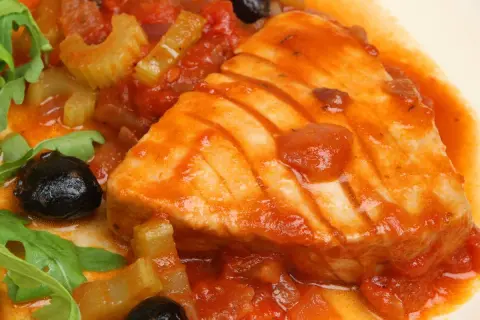 Un succulento tonno in padella con pomodoro, olive nere e sedano, guarnito con rucola fresca.