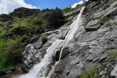 Una hermosa cascada en Madrid, cayendo por rocas grises y vegetación verde bajo un cielo azul con nubes blancas.