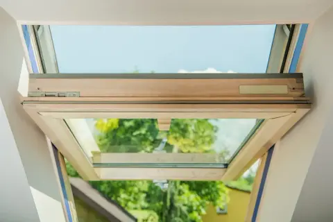 Ventana de tejado abierta, mostrando el marco de madera y el cristal con cámara de aire. Se ve el cielo azul y árboles verdes al exterior.