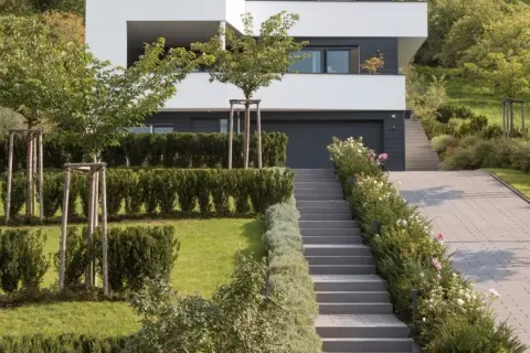 Moderner Garten am Hang mit gepflasterter Auffahrt, Treppen und gepflegten Hecken. Ein Haus mit weißen Fassaden und dunklen Fenstern dominiert den Hintergrund.