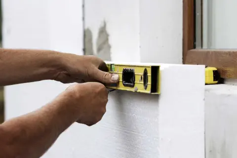 Ręce budowlańca sprawdzają poziomicą, jakie są grubości styropianu przy oknie.