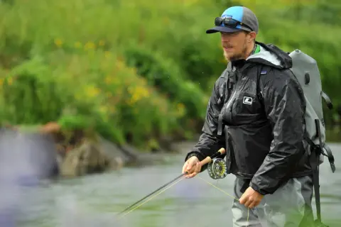 Ein Mann mit Hut und Sonnenbrille steht im Wasser und übt das **streamer fischen**. Er trägt eine schwarze Regenjacke und einen Rucksack.