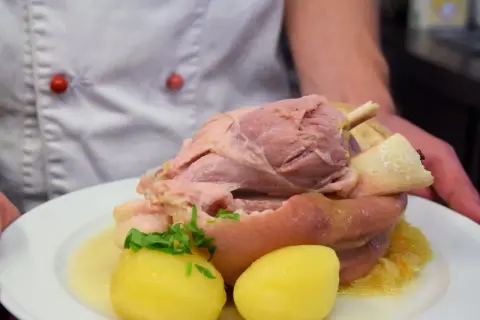 Ein Koch präsentiert ein deftiges Eisbein mit Kartoffeln und Sauerkraut. Die Frage "Eisbein wie lange kochen?" beschäftigt viele Hobbyköche.