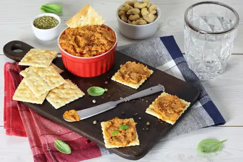 Pyszna pasta z suszonych pomidorów na krakersach, idealna na przekąskę. Obok miseczka z orzechami i szklanka wody.