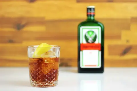 Orzeźwiający jager z colą, z plasterkiem cytryny, obok zielonej butelki Jägermeistera.